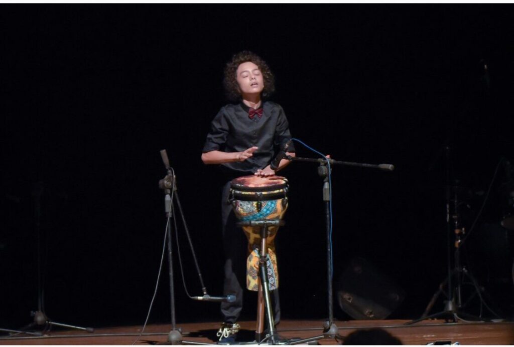 Aurum playing the djembe in black on a dark stage